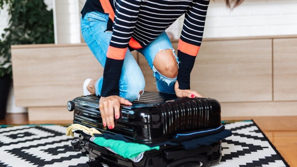 Woman at home on the floor struggling to close her overpacked luggage.
