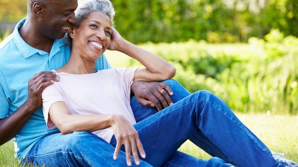 Moving abroad at age 50. A couple is sitting on the grass. They are both wearing blue jeans. He is wearing a light blue shirt and she is wearing a light pink shirt. He is holding her while they are both smiling.