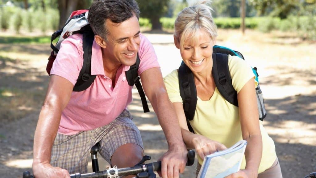 A couple is riding bicycles on a trail. The stop to look at directions on a map. He is wearing a pink shirt and plaid shorts with a small backpack on his back. She is wearing a yellow shirt, and khaki shorts. She is also wearing a small backpack.