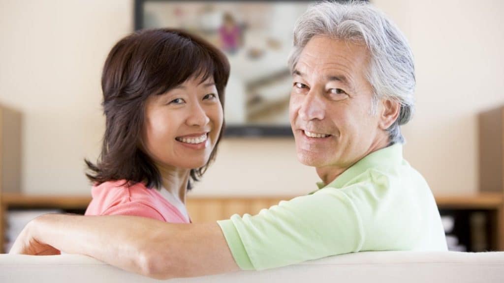 A couple sitting on couch smiling and looking at Television together. She has dark hair and she is wearing a pink shirt. He has his arm around her and he is wearing a light green shirt.