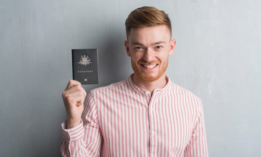 Man with red hair and beard is wearing a red and white striped, long sleeved shirt. He is holding up a Passport with his right hand and smiling.