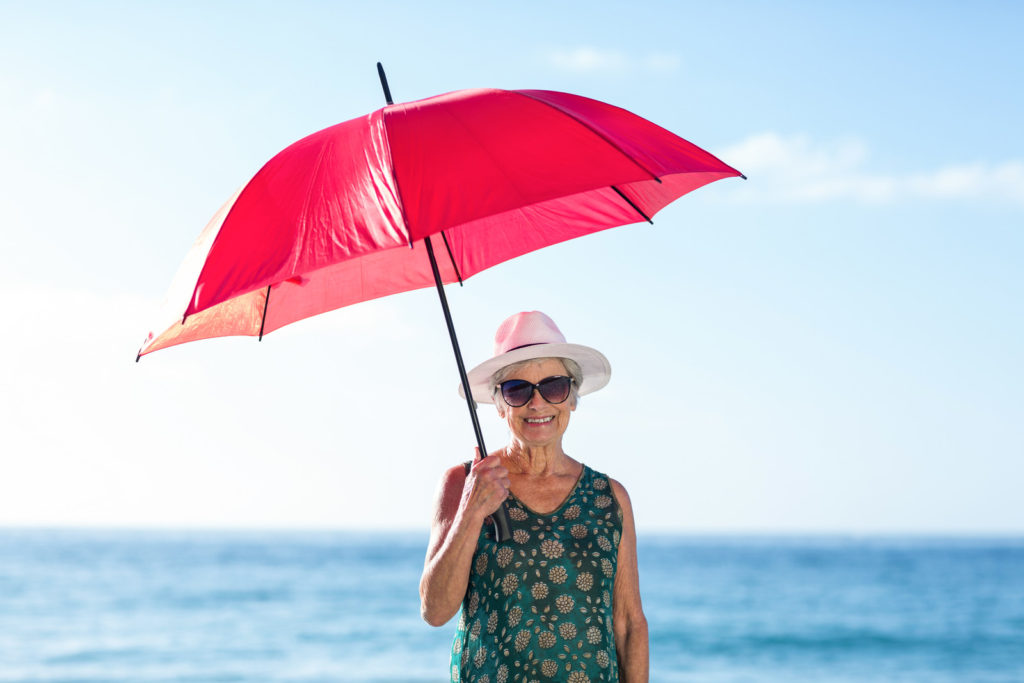 A lady is holding a red umbrella. She is wearing a green dress and a white hat. She is wearing shades and smiling. The Sea is in the background.