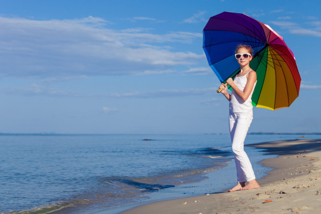A girl with a colorful umbrella is wearing white pants, white tank top, and white shades. She is standing on a beach.