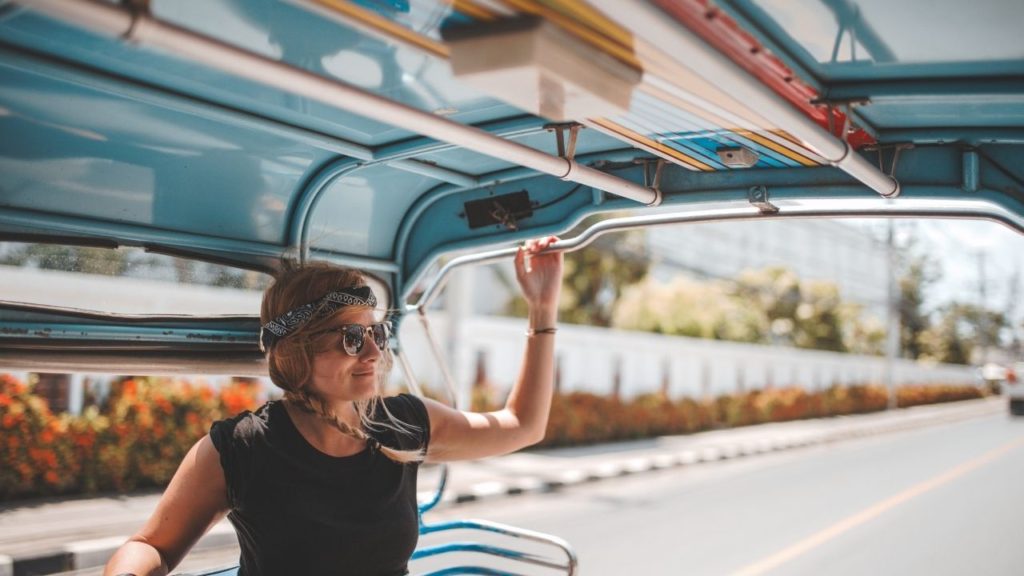 A woman rides in the back of a taxi or truck transport. She is wearing shades and a black top. Her hair is braided with a headband around her hair.