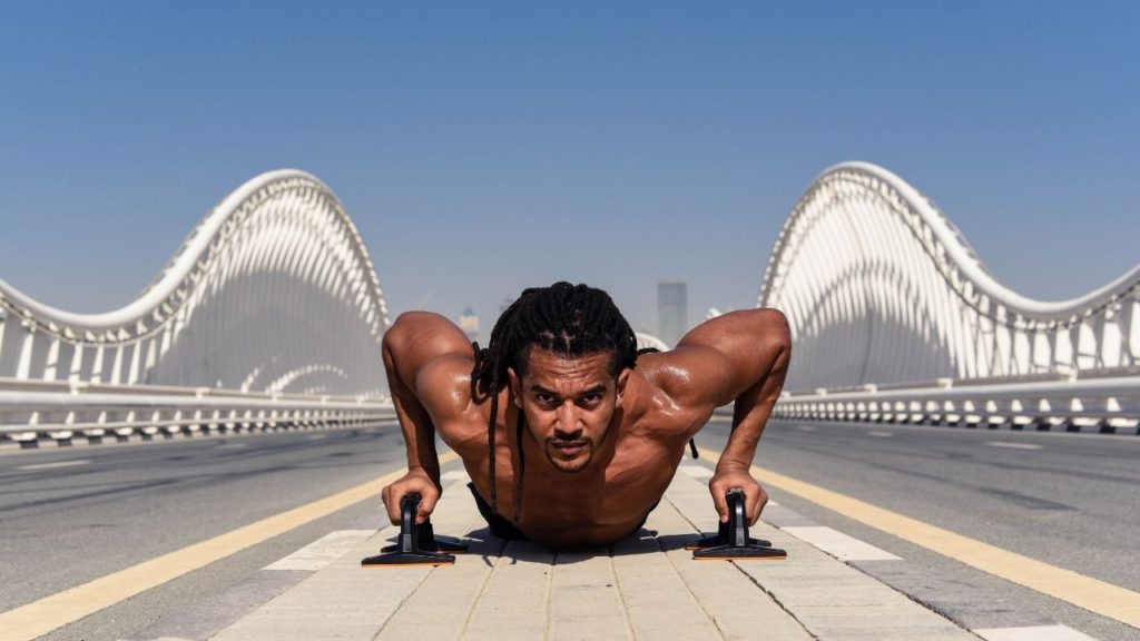 Black man with dreadlocks, and no shirt on is exercising on a white bridge.