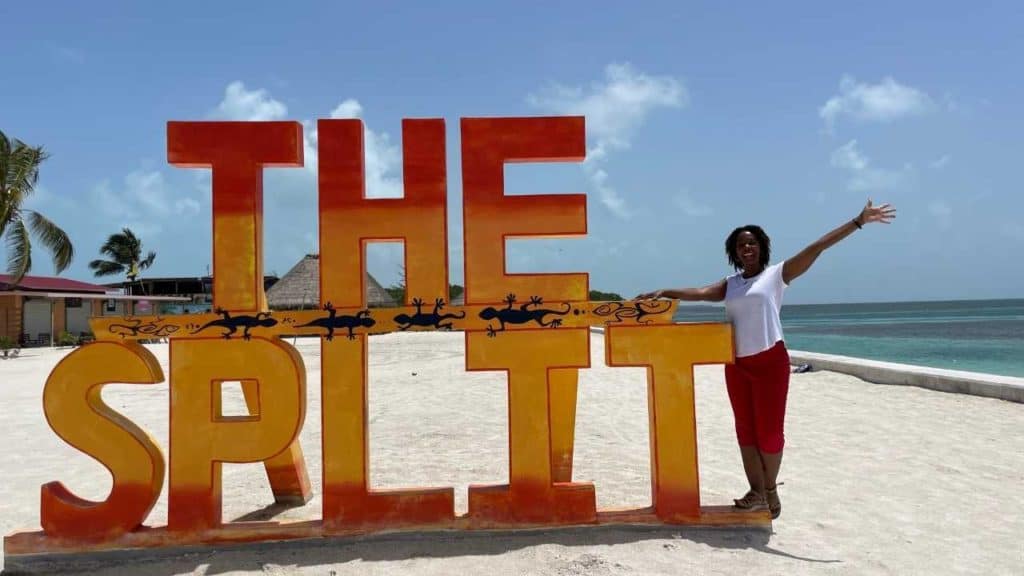 Kedra at The Split. She is wearing white top and red shorts. The split sign is orange and yellow with beach sand and the ocean in the background.